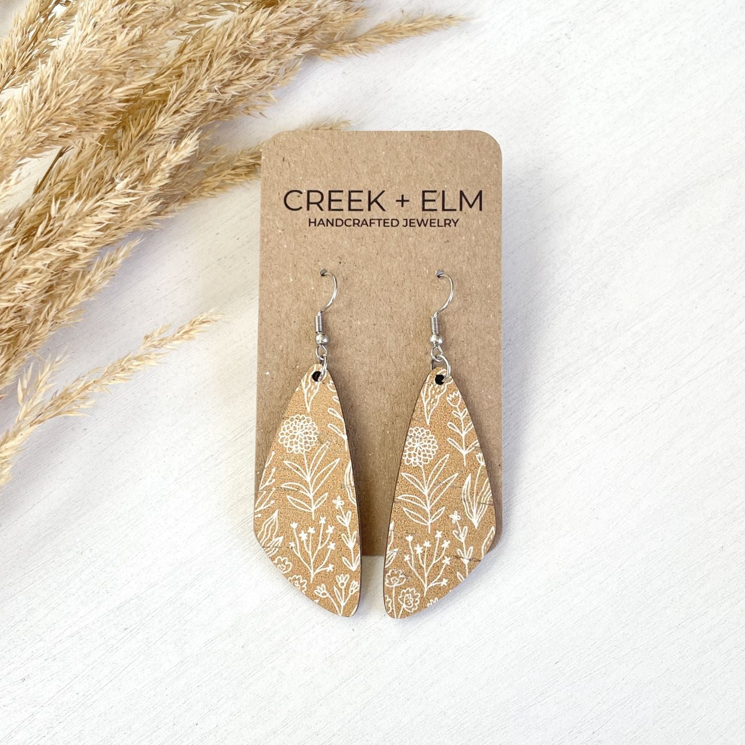 A pair of teardrop-shaped dangle earrings with a beige and white botanical pattern, displayed on a brown kraft card labeled 'CREEK + ELM HANDCRAFTED JEWELRY' on a white textured background with dried grass.