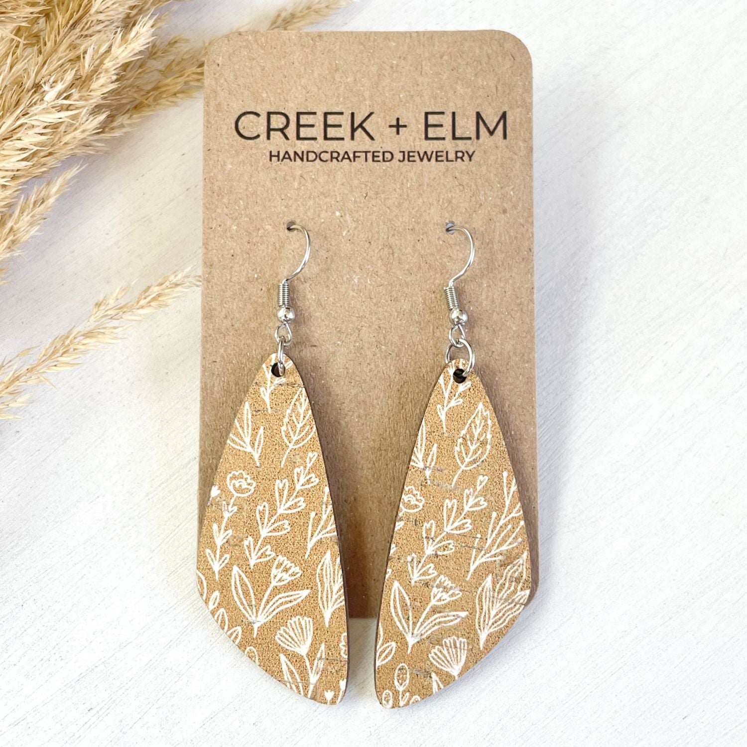 A pair of teardrop-shaped dangle earrings with a tan and white botanical pattern, displayed on a brown kraft card labeled 'CREEK + ELM HANDCRAFTED JEWELRY' on a white background with dried grass.