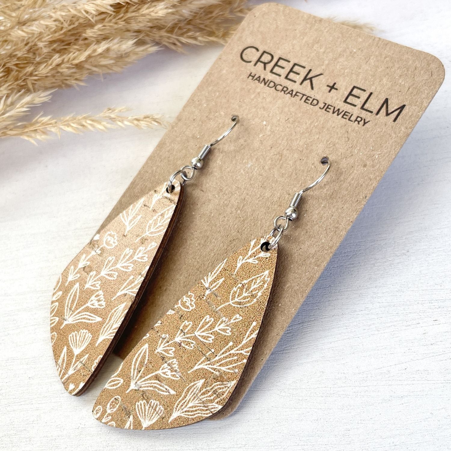 Teardrop-shaped wooden earrings with white leaf and floral line patterns, displayed on a brown kraft card labeled 'CREEK + ELM HANDCRAFTED JEWELRY' on a light wood surface with dried grass in the background.