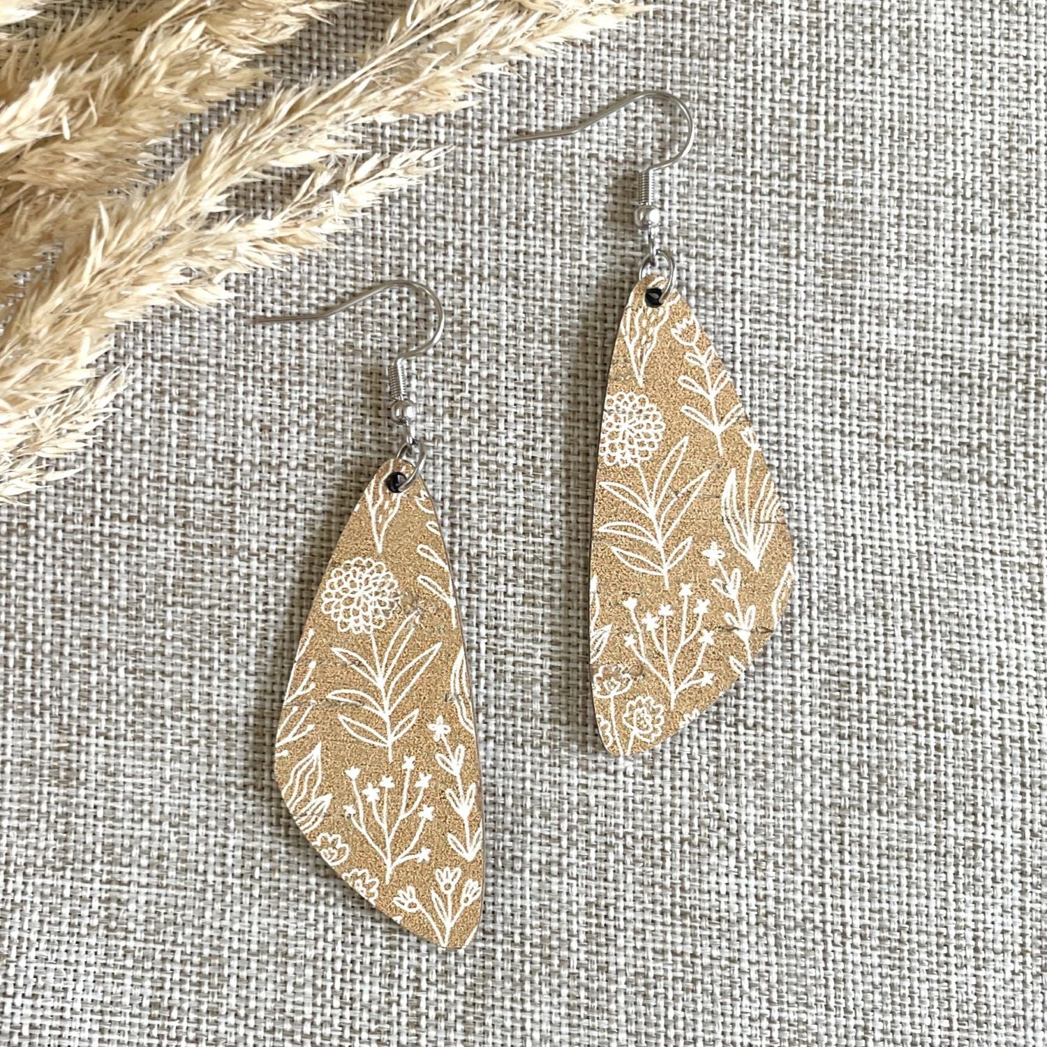 A pair of teardrop-shaped dangle earrings with a tan and white botanical pattern, displayed on a textured beige fabric background with dried grass as a prop.