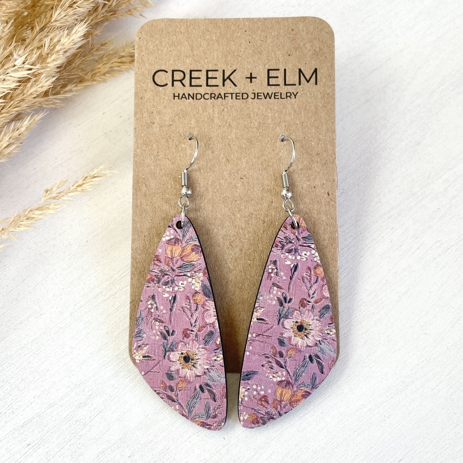 A pair of teardrop-shaped dangling earrings with a pink floral pattern, displayed on a brown kraft card labeled CREEK + ELM HANDCRAFTED JEWELRY. The earrings are photographed on a light textured surface with dried grass as a decorative prop.