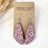 A pair of teardrop-shaped dangling earrings with a pink floral pattern, displayed on a brown kraft card labeled CREEK + ELM HANDCRAFTED JEWELRY. The earrings are photographed on a light textured surface with dried grass as a decorative prop.
