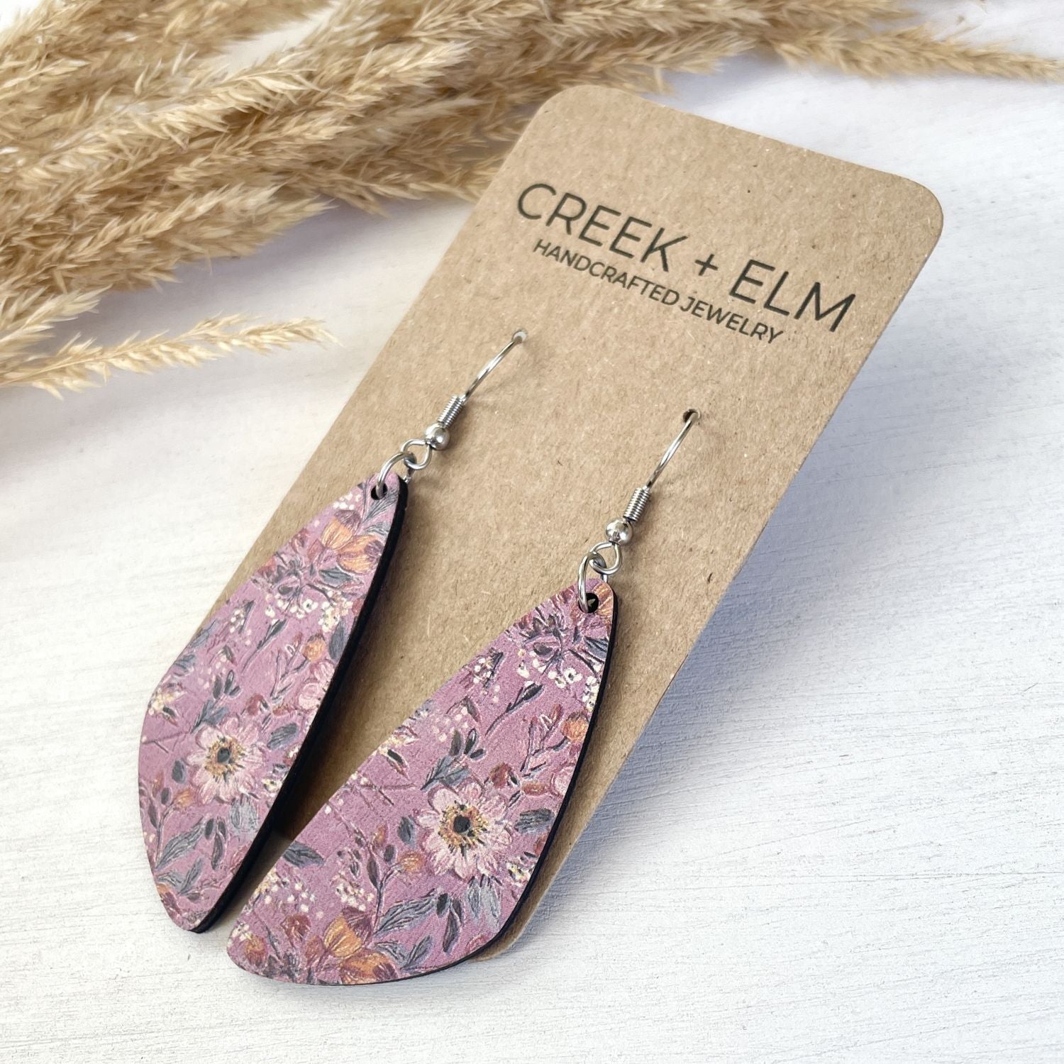 Pair of teardrop-shaped dangling earrings with a dusty pink floral pattern, displayed on a brown kraft card labeled CREEK + ELM. The earrings are photographed on a white wooden surface with dried grass in the background.