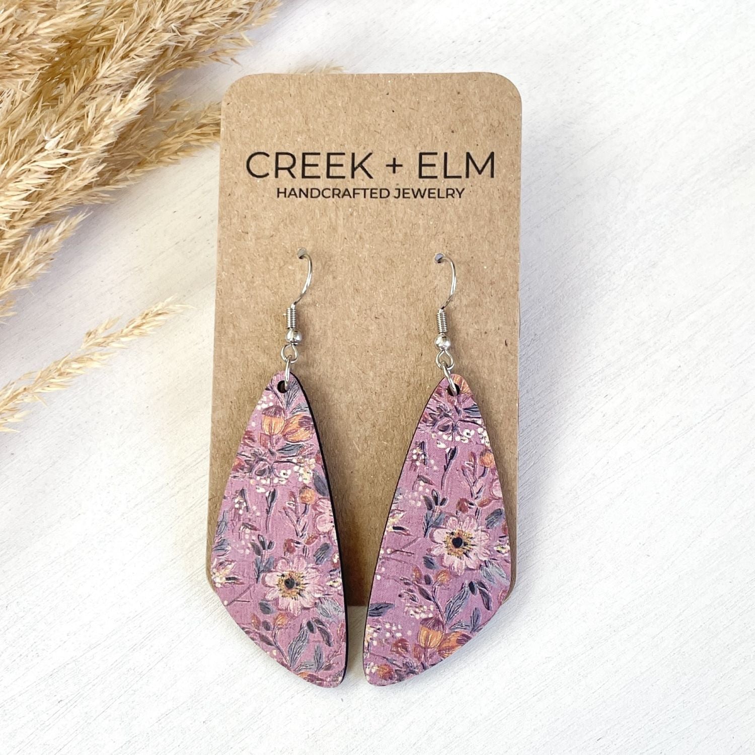 Teardrop-shaped dangle earrings with a pink floral pattern, displayed on a brown kraft card labeled 'CREEK + ELM HANDCRAFTED JEWELRY' on a white textured background with dried grass accents.