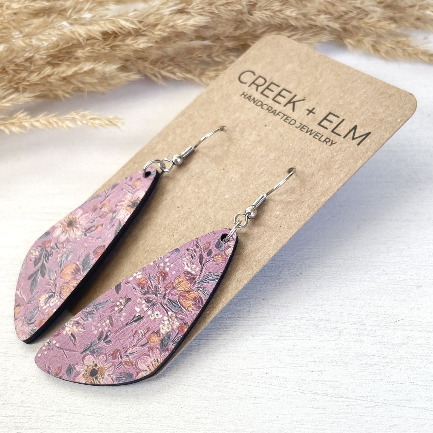 Teardrop-shaped dangle earrings with a dusty pink floral pattern, displayed on a brown kraft card labeled 'CREEK + ELM HANDCRAFTED JEWELRY' on a white wooden surface with dried pampas grass in the background.