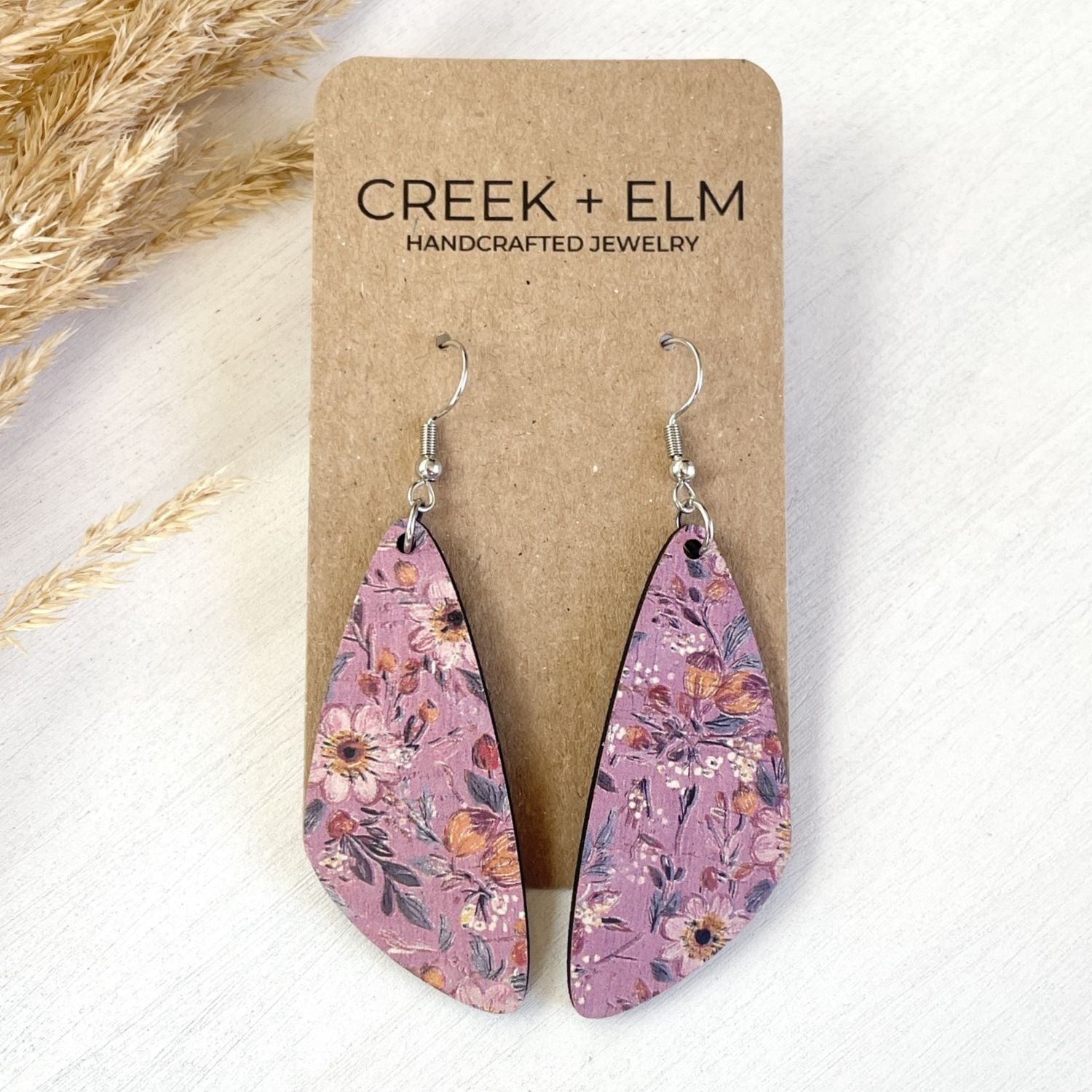 A pair of teardrop-shaped dangling earrings with a floral print on a kraft display card. The card reads 'CREEK + ELM HANDCRAFTED JEWELRY.