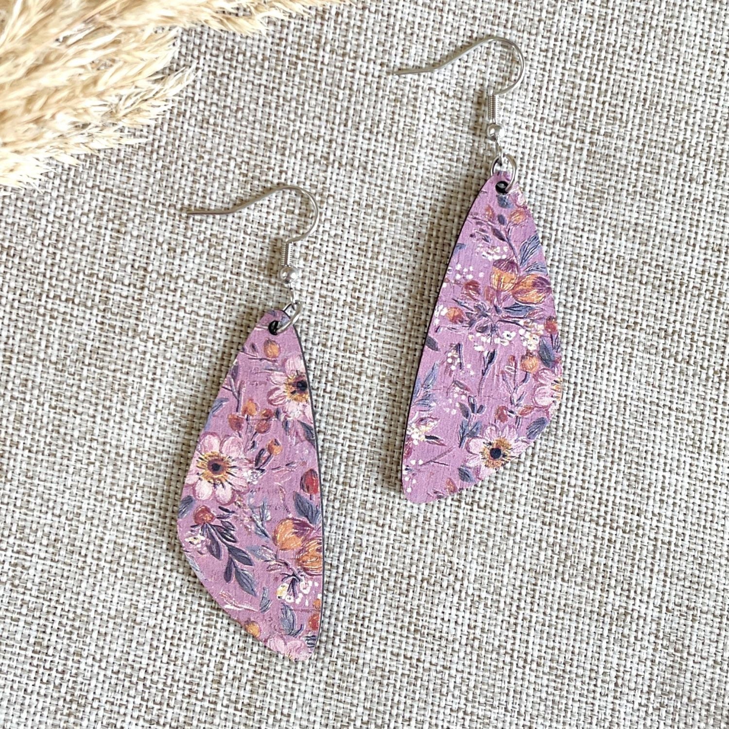 A pair of teardrop-shaped dangle earrings with a pink floral pattern, hanging from silver fishhook wires on a textured beige fabric background.