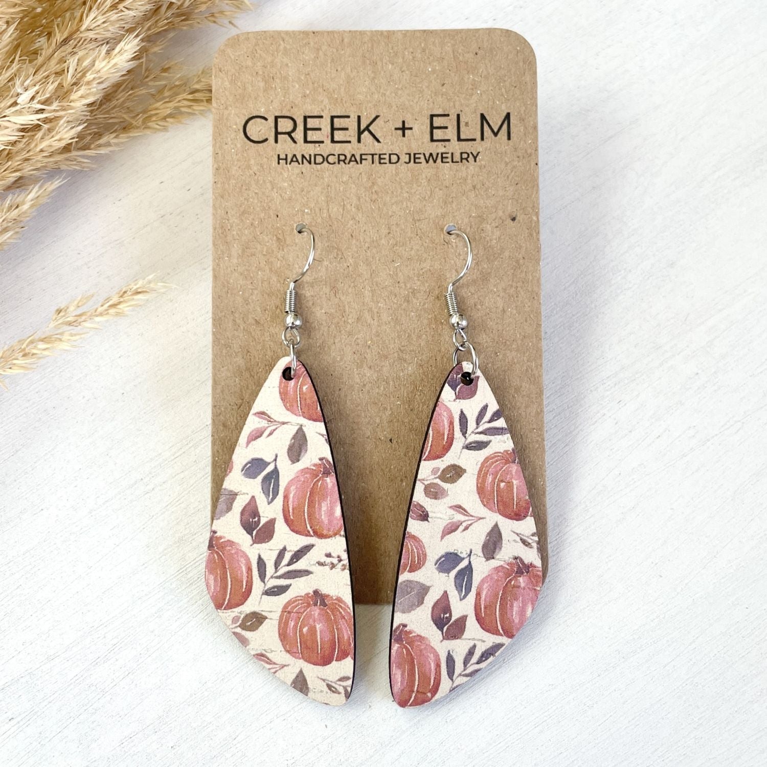Teardrop-shaped dangle earrings with a fall harvest pumpkin pattern, displayed on a brown kraft card labeled 'CREEK + ELM HANDCRAFTED JEWELRY' on a white background with dried grass accents.