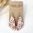 Teardrop-shaped dangle earrings with a fall harvest pumpkin pattern, displayed on a brown kraft card labeled 'CREEK + ELM HANDCRAFTED JEWELRY' on a white background with dried grass accents.