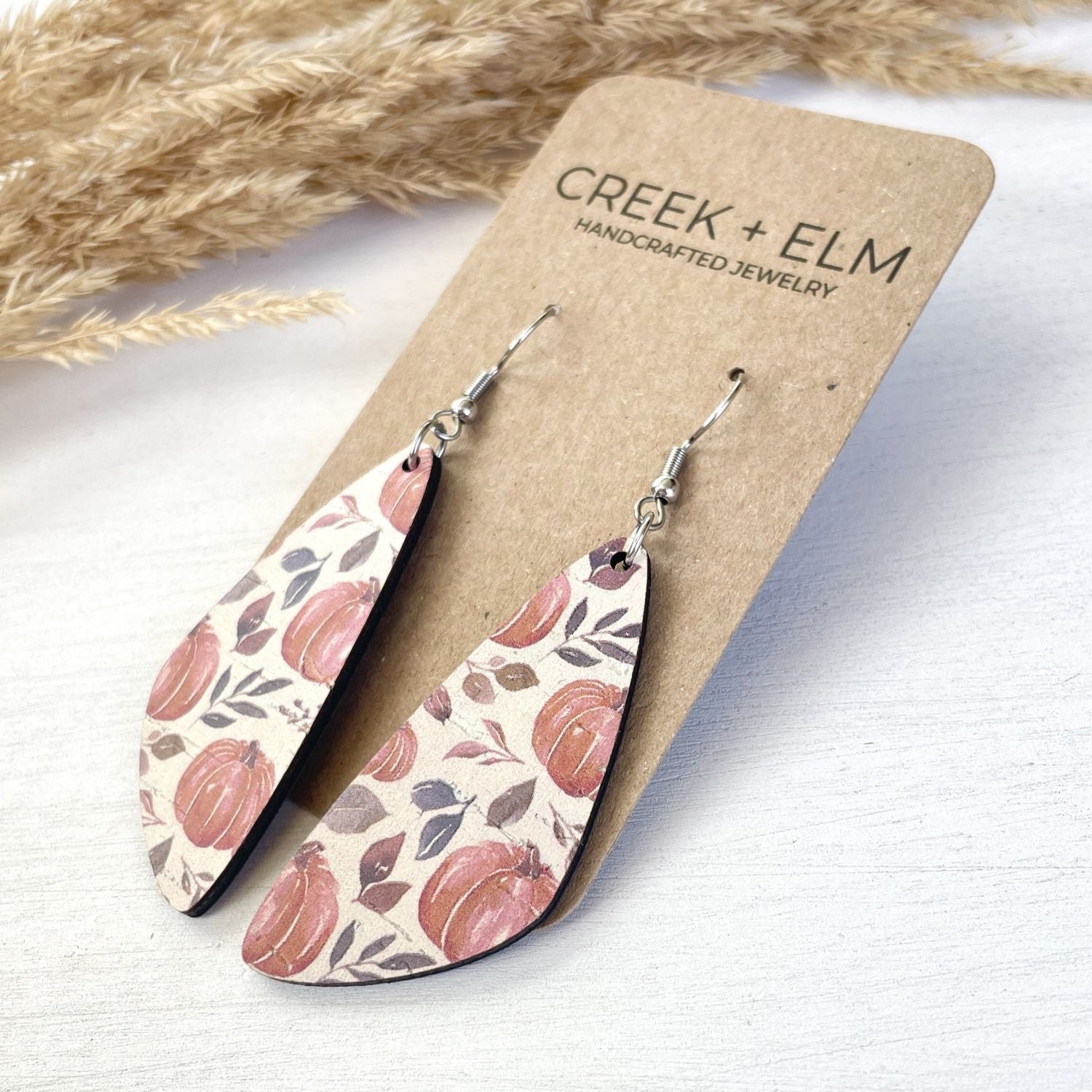 A pair of teardrop-shaped dangle earrings with a fall pumpkin and leaf pattern, displayed on a brown kraft card labeled 'CREEK + ELM HANDCRAFTED JEWELRY'. The earrings are photographed on a light-colored wooden surface with dried pampas grass in the background.