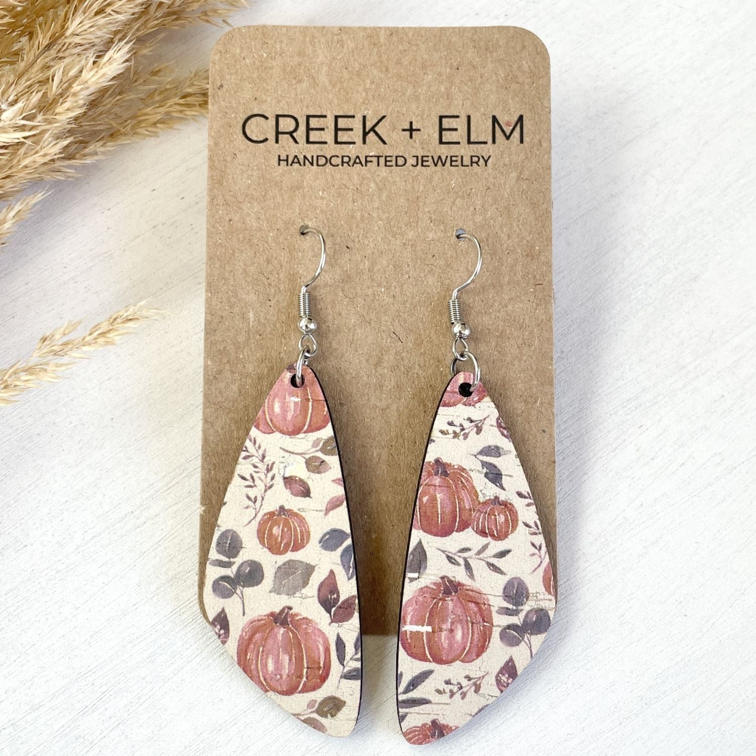 A pair of teardrop-shaped dangle earrings with a fall harvest pattern of pumpkins, leaves, and berries on a cream background, hanging from silver hooks on a brown cardboard display card labeled 'CREEK + ELM HANDCRAFTED JEWELRY'.