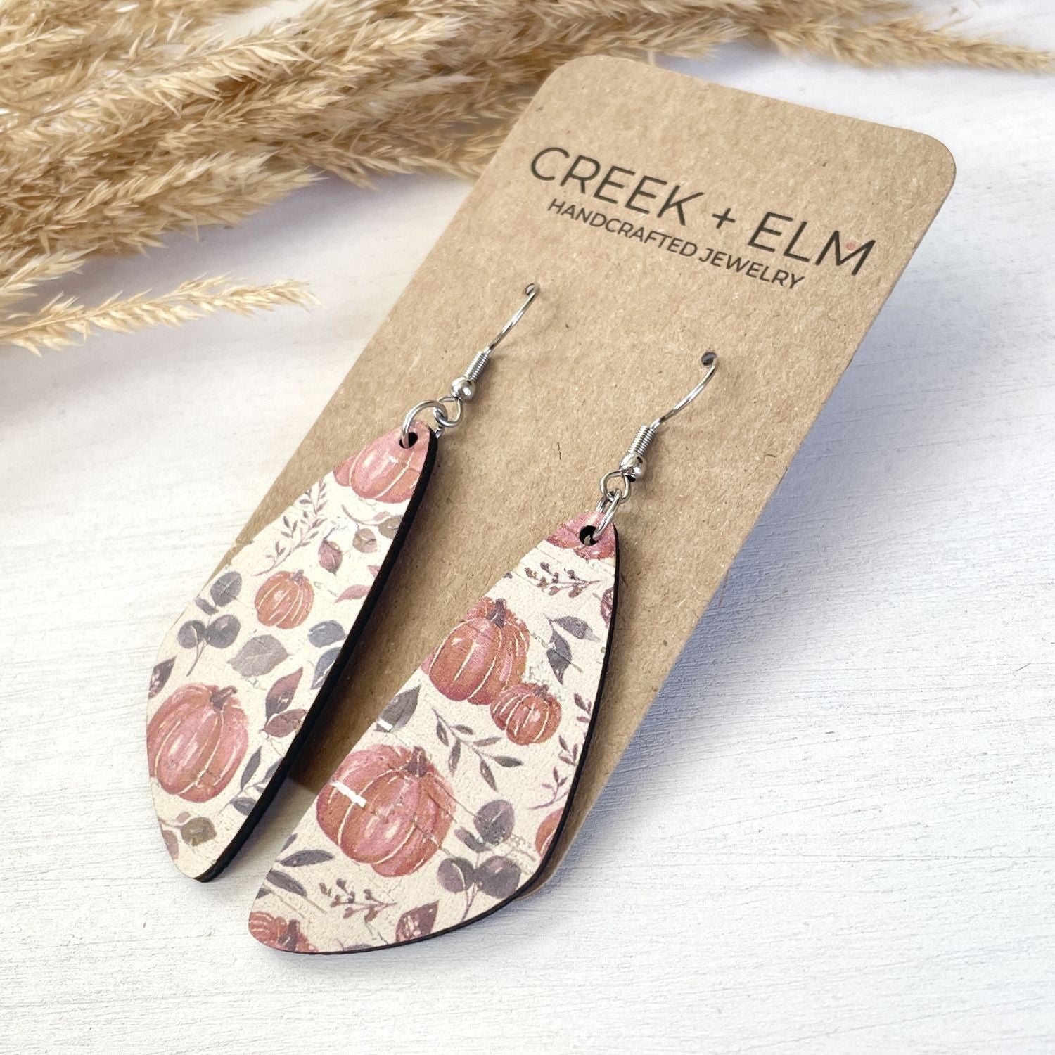 A pair of teardrop-shaped dangle earrings with a fall pumpkin pattern, displayed on a brown kraft card labeled 'CREEK + ELM'.
