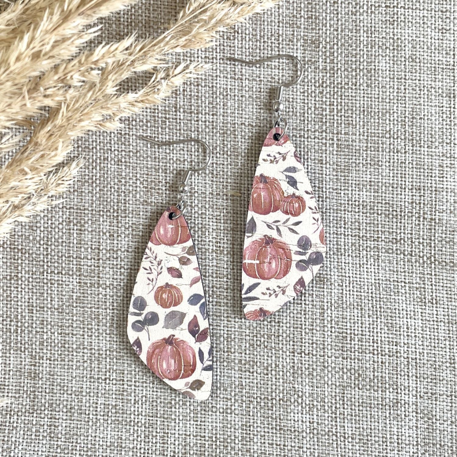 A pair of irregularly shaped dangle earrings with a fall harvest pattern of pumpkins, leaves, and berries on a cream background, hanging from silver fishhook wires. The earrings are displayed on a textured beige linen fabric with dried wheat stalks in the top-left corner.