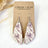 A pair of teardrop-shaped dangle earrings with a vintage witch illustration pattern in purple and brown on a cream background, hanging from silver hooks on a brown kraft display card labeled CREEK + ELM.