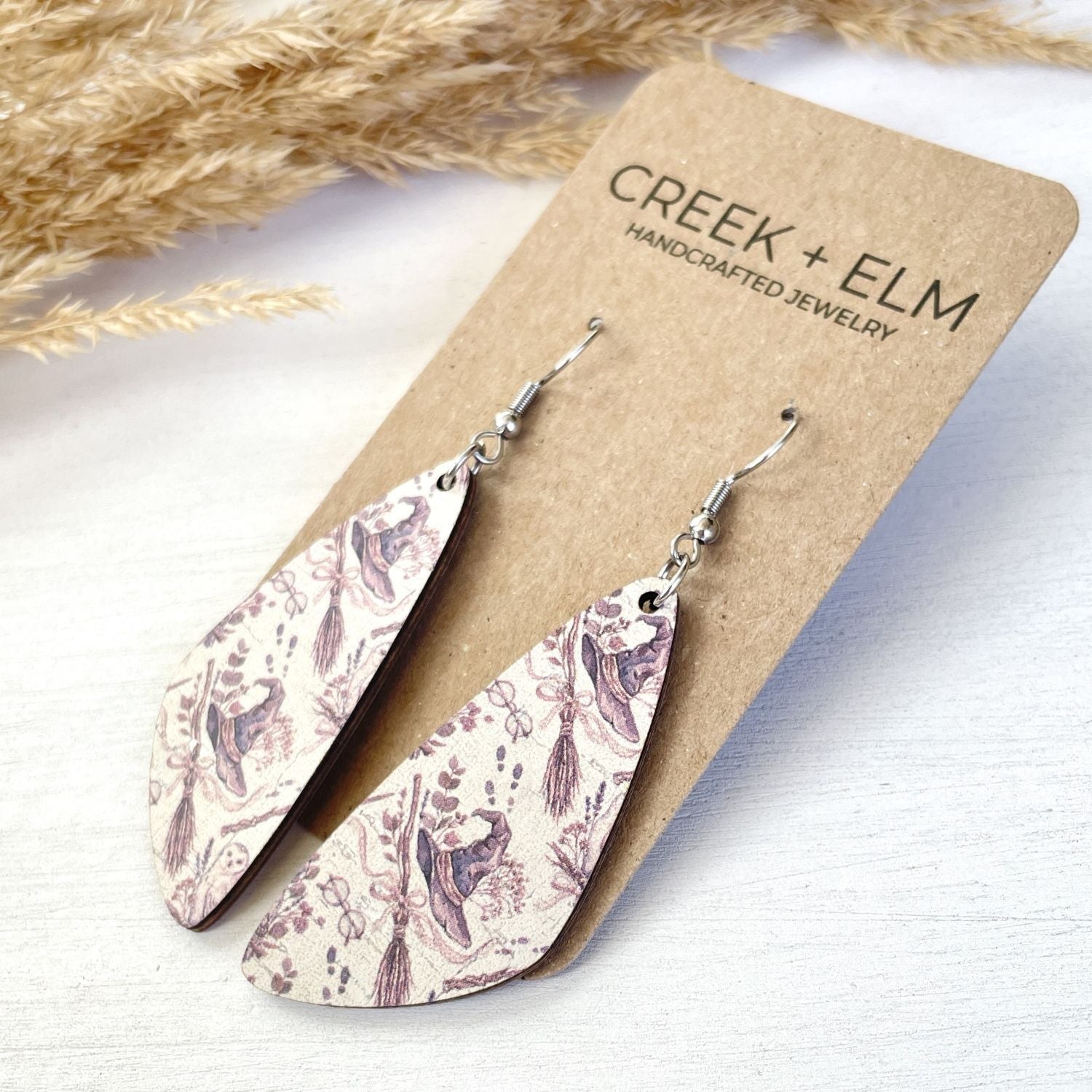 Teardrop-shaped dangle earrings with a purple and beige floral pattern, displayed on a brown kraft card labeled CREEK + ELM. The card is set against a light wood background with dried grass in the upper left corner.