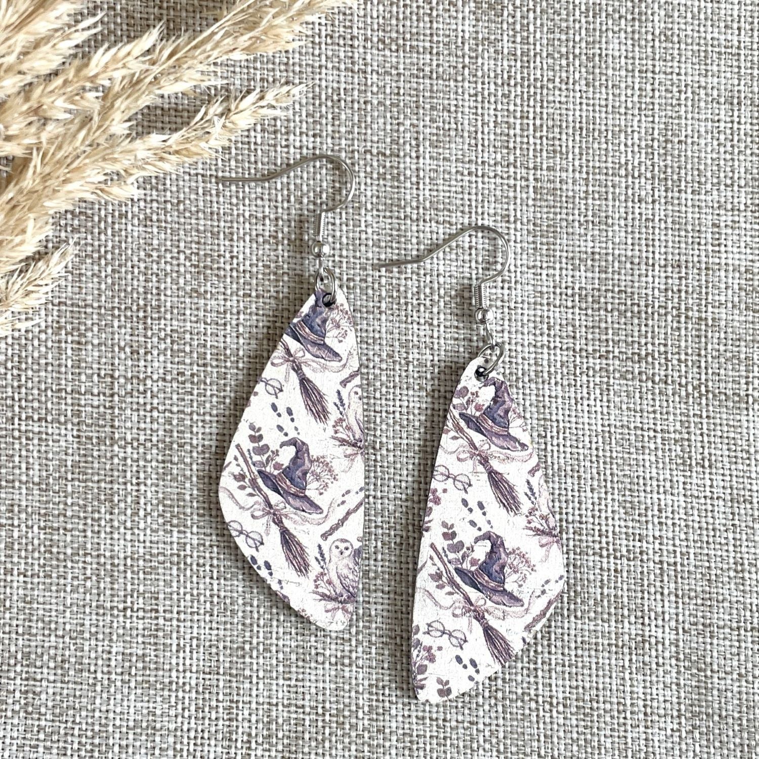 A pair of teardrop-shaped dangle earrings with a white background and a purple botanical illustration pattern, hanging on silver fishhook wires. The earrings are displayed on a textured beige linen fabric background with dried grass in the top-left corner.