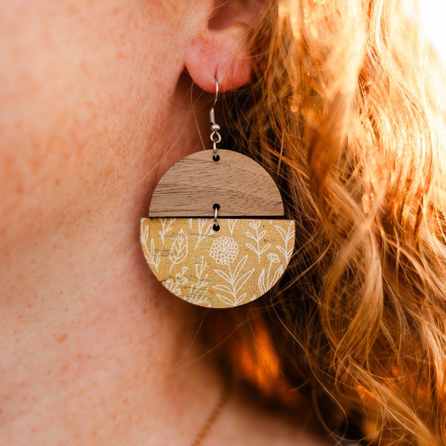 A close-up of a person's ear wearing a large circular drop earring. The earring is split horizontally into two halves: the top half is natural wood, and the bottom half features a yellow and white botanical patterned print.