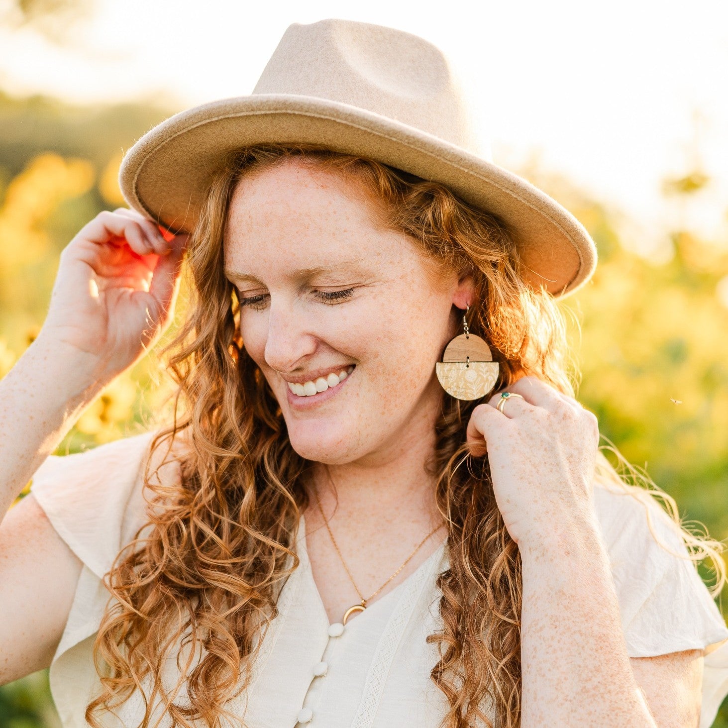 A smiling woman with long curly red hair wearing a beige wide-brim hat and matching beige top, holding her hair and wearing a large circular wooden earring in a sunlit outdoor setting.