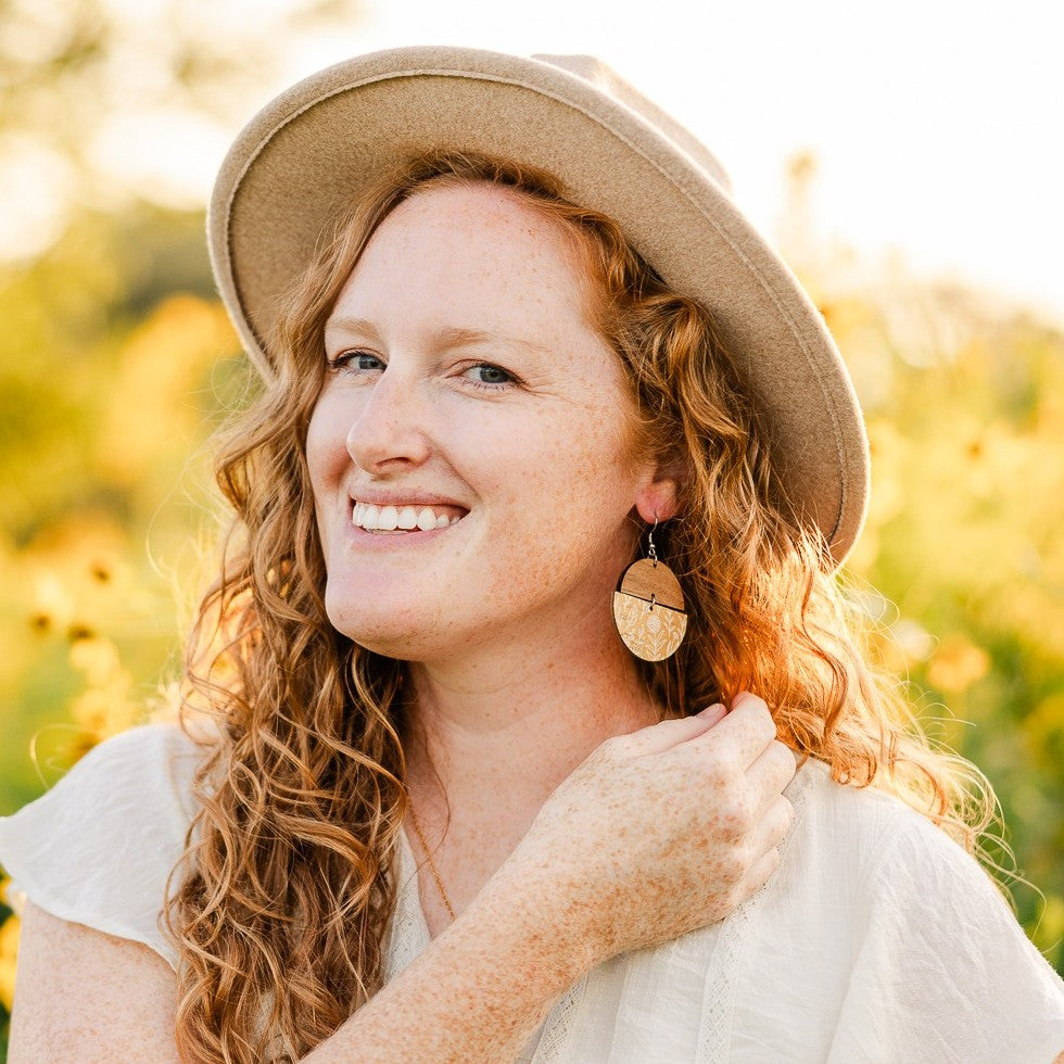 A smiling woman with curly red hair wearing a beige wide-brim hat and a white top, photographed outdoors in warm golden light with blurred foliage in the background.