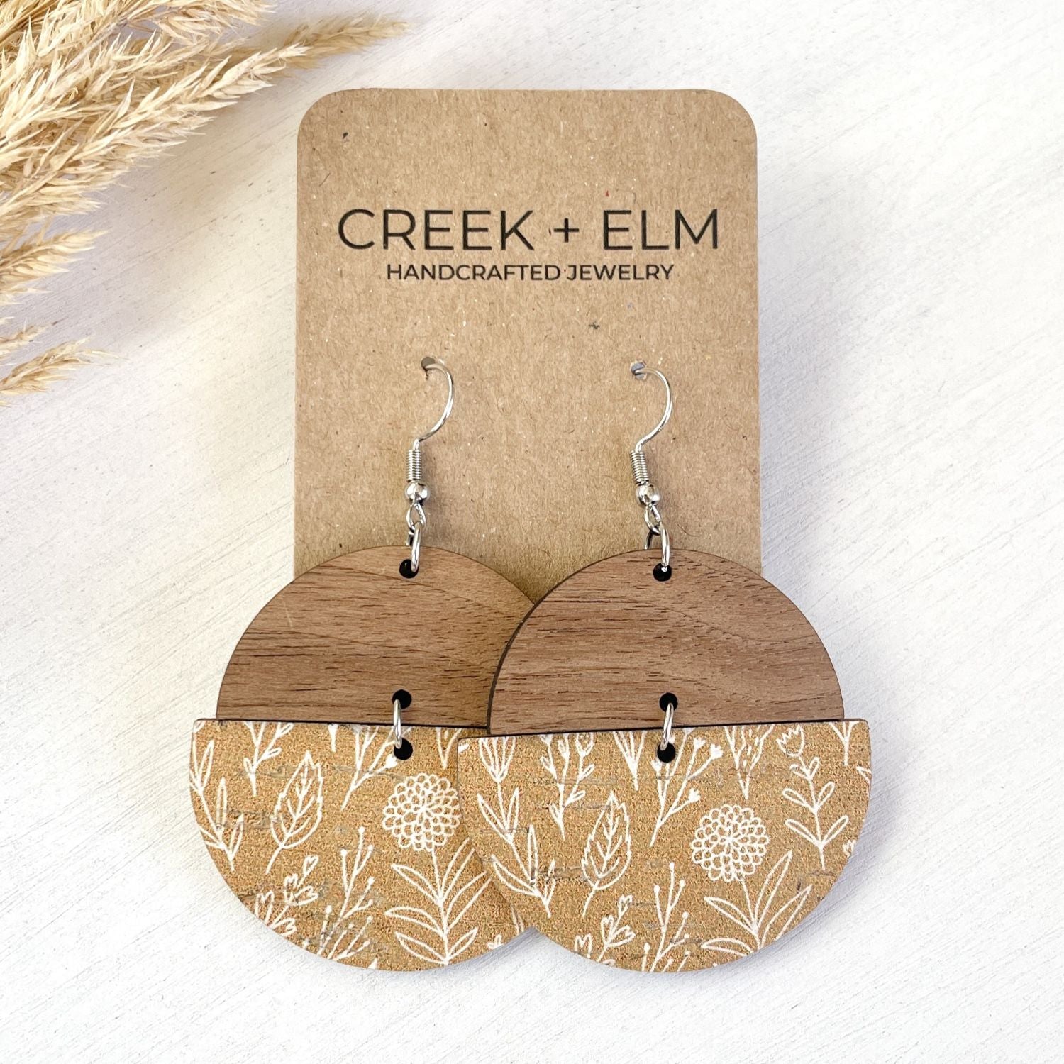 A pair of handmade dangle earrings displayed on a kraft paper card. The earrings feature a warm brown wood top half and a tan fabric bottom half with a white botanical leaf pattern.