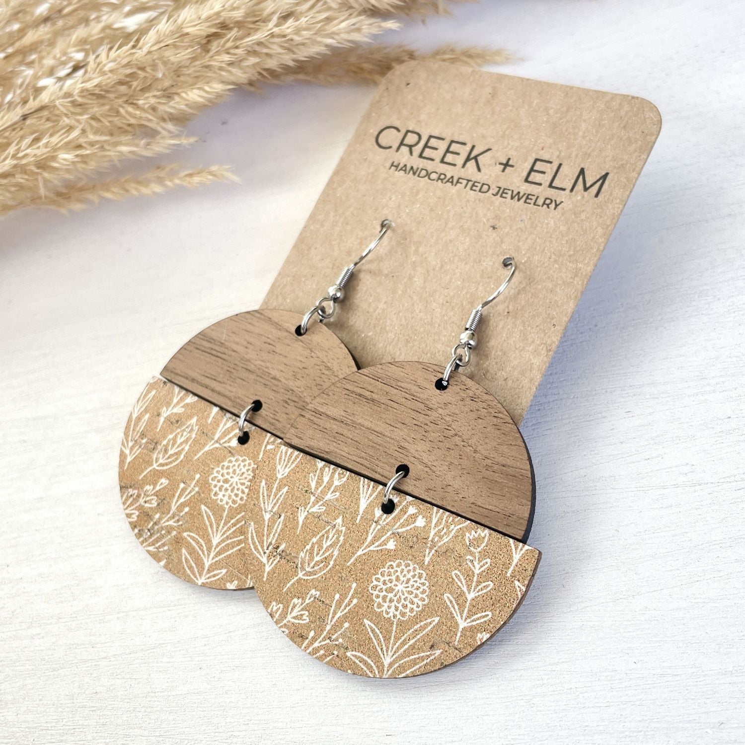 Wooden dangle earrings with a natural wood top half and a floral patterned lower half, displayed on a kraft card labeled CREEK + ELM HANDCRAFTED JEWELRY on a light wood surface.