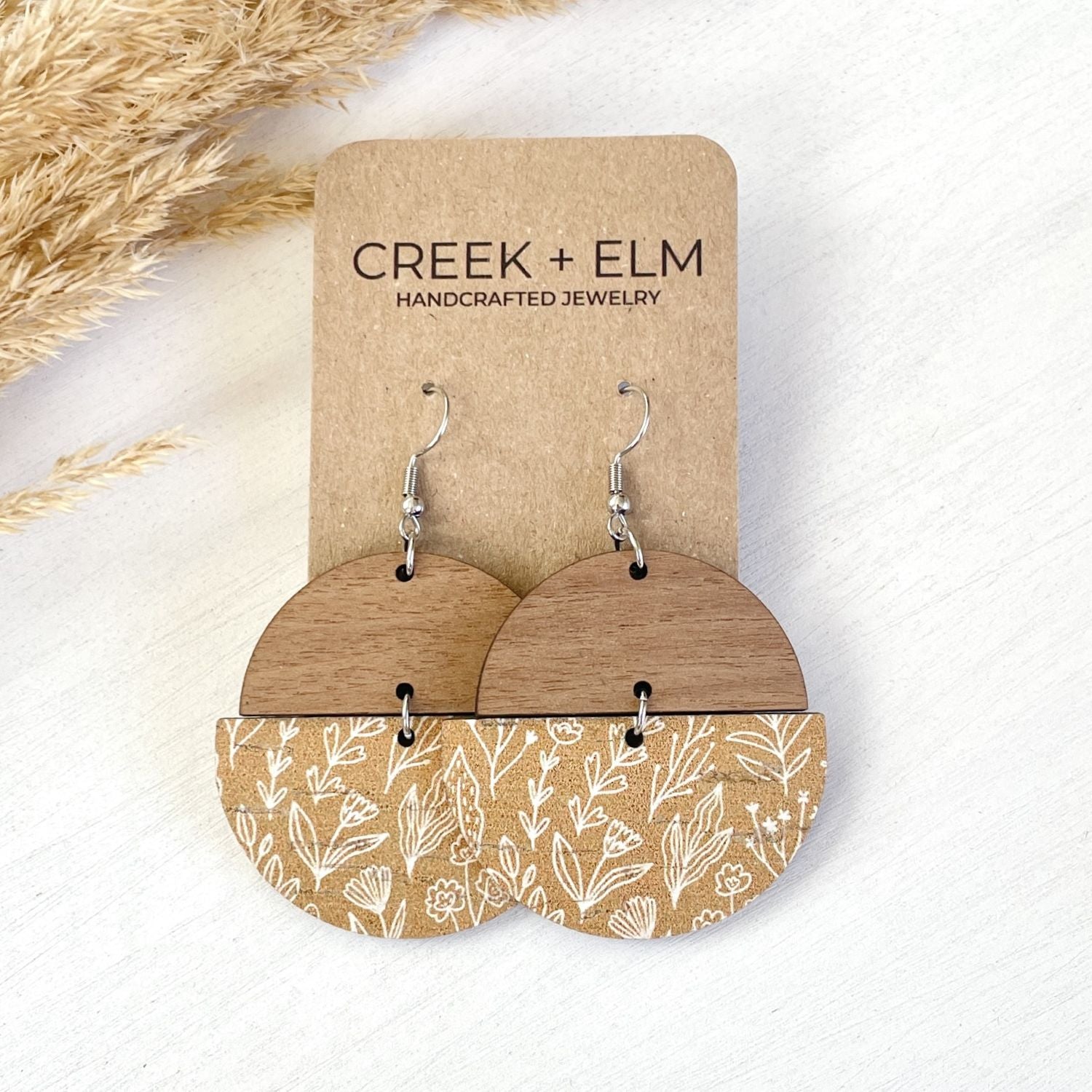 Wooden dangle earrings displayed on a kraft card. The earrings feature a solid wood top half and a lower half with a white botanical line-art pattern on a tan wood base.