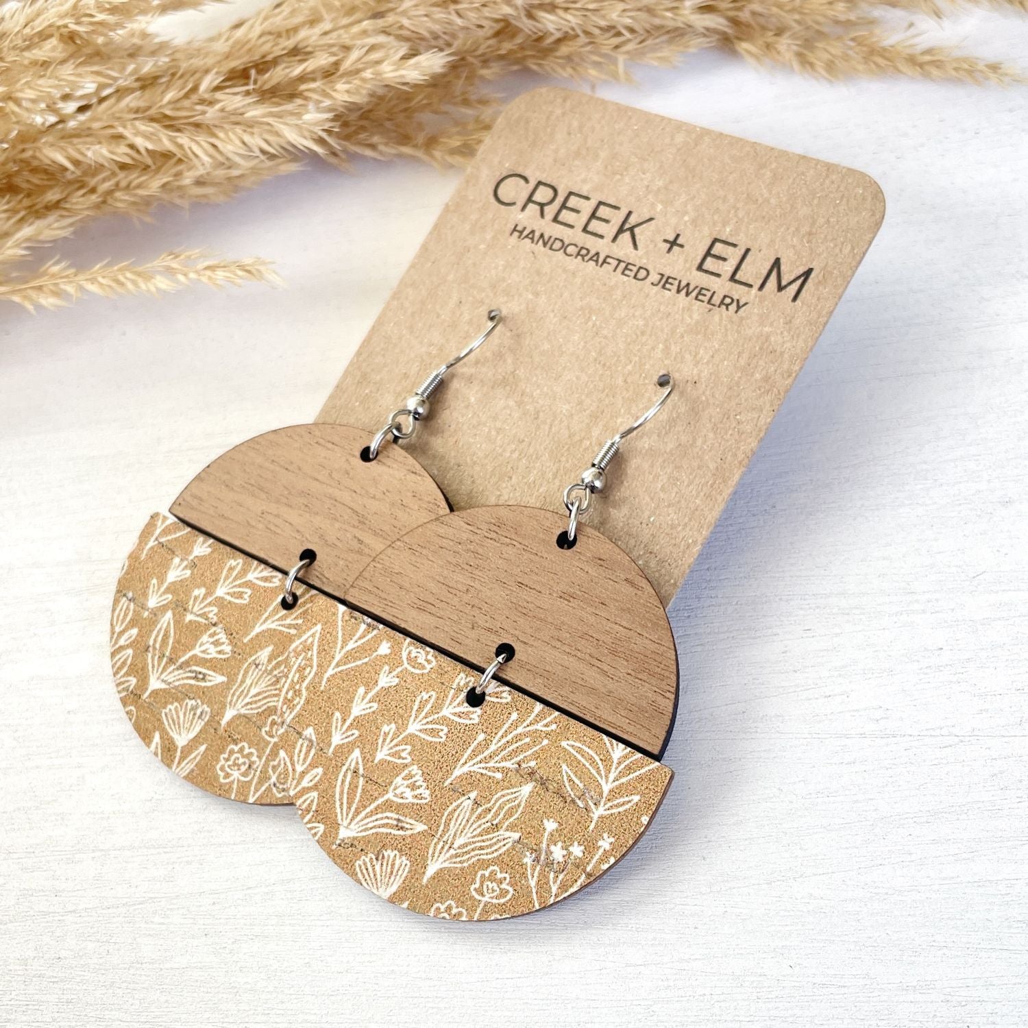 Pair of wooden dangle earrings with a natural wood top half and a printed tan-and-white floral bottom half, hanging from silver hooks on a kraft display card that reads CREEK + ELM HANDCRAFTED JEWELRY.