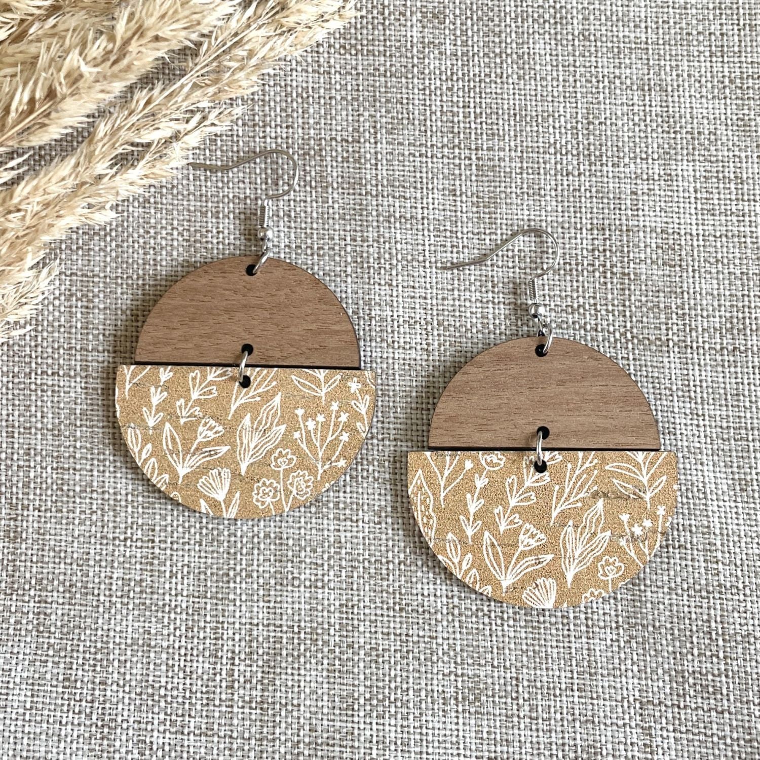 A pair of handmade dangle earrings with a split design: the top half is natural wood, and the bottom half has a beige floral pattern. They hang from silver fishhook wires on a textured beige fabric background with dried grass in the top-left corner.