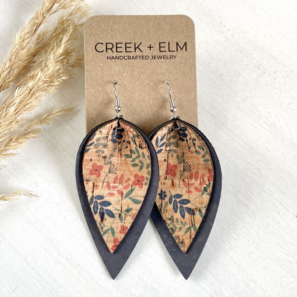 Teardrop-shaped earrings with a floral patterned center and dark charcoal outer edges, hanging from silver hooks on a kraft display card. The card reads 'CREEK + ELM HANDCRAFTED JEWELRY' and is styled on a white surface with dried grass.