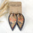 Teardrop-shaped earrings with a floral patterned center and dark charcoal outer edges, hanging from silver hooks on a kraft display card. The card reads 'CREEK + ELM HANDCRAFTED JEWELRY' and is styled on a white surface with dried grass.