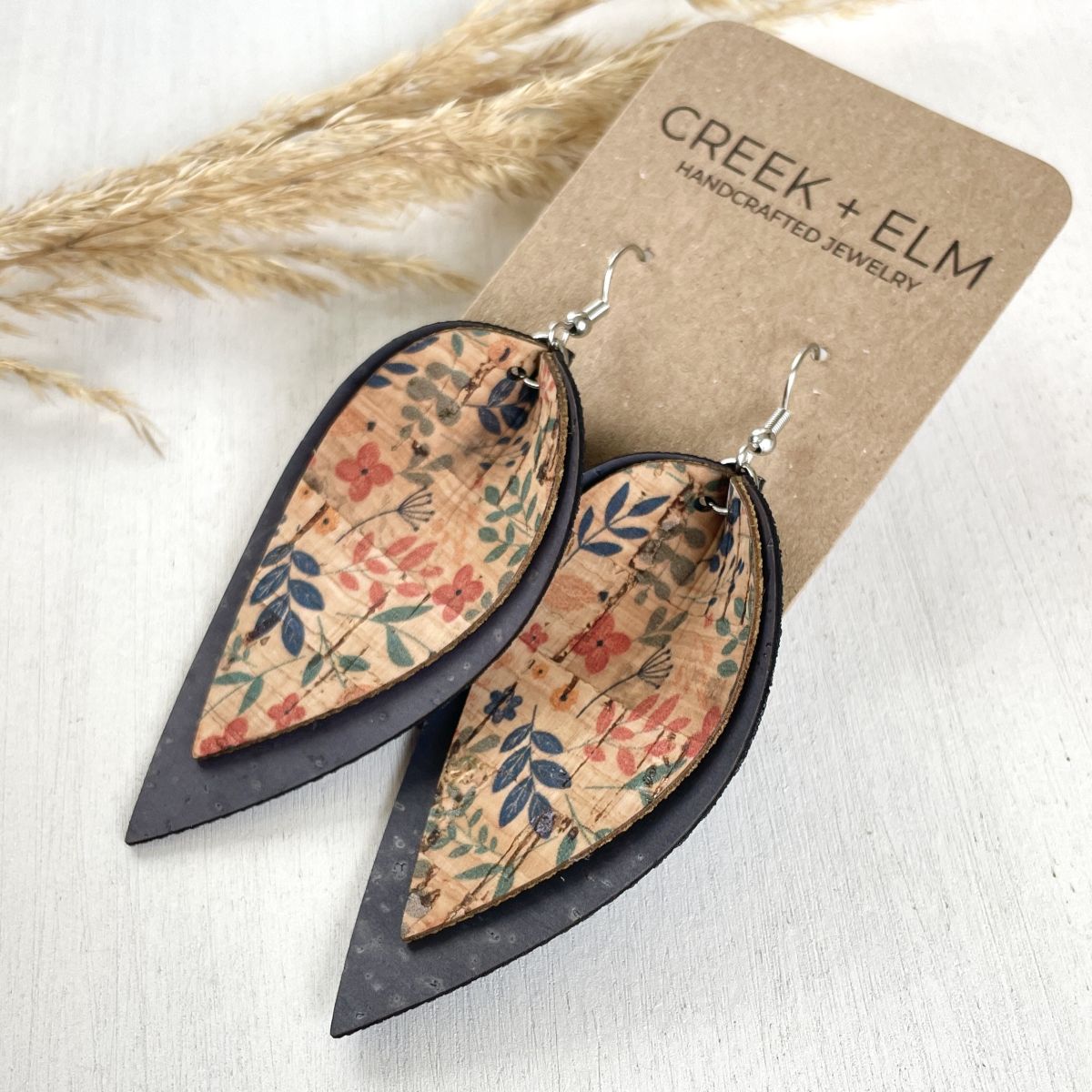 Teardrop-shaped dangle earrings with a floral patterned center and dark outer border, displayed on a brown cardboard card labeled CREEK + ELM. The earrings are photographed on a light surface with dried grass as a styled prop.