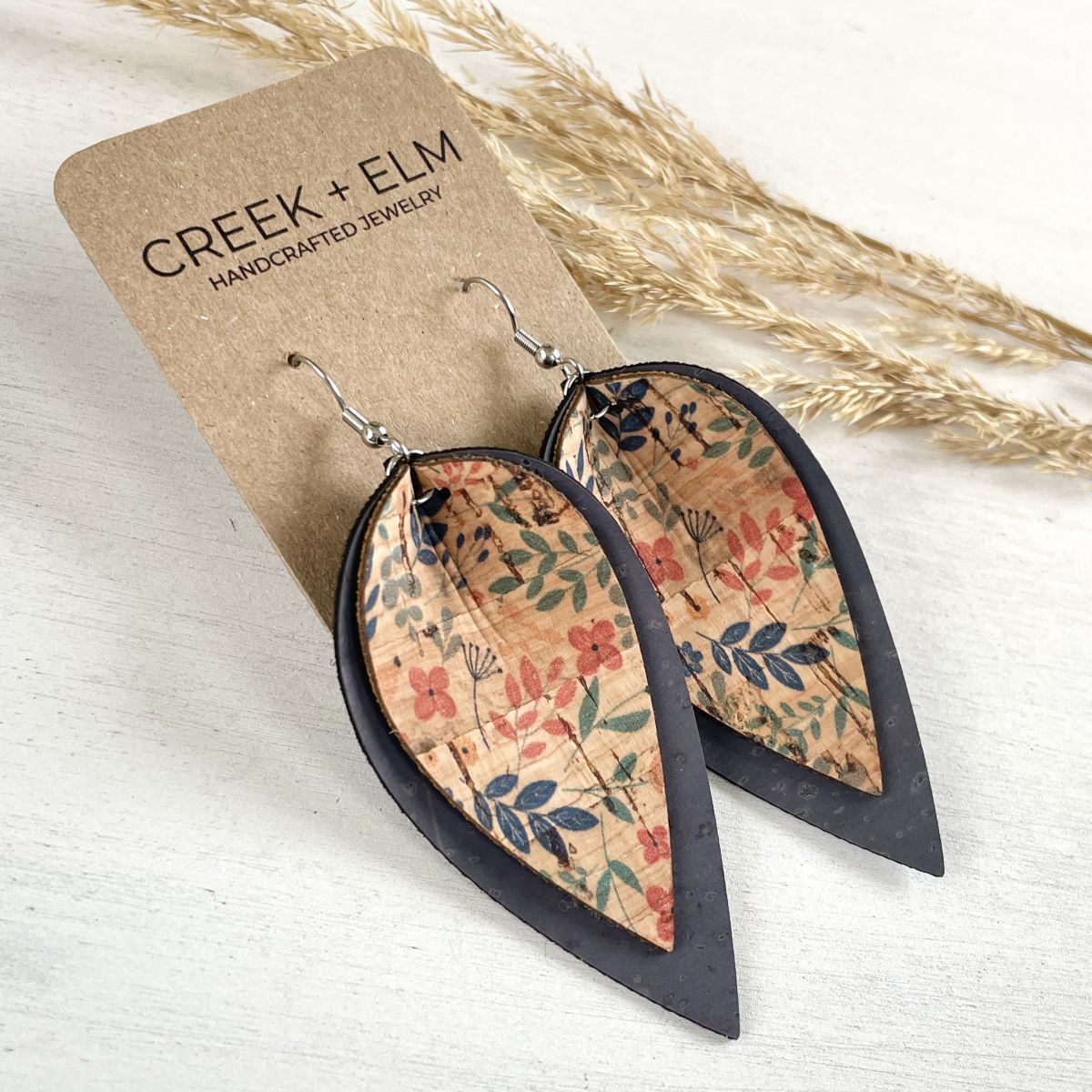 Teardrop-shaped dangle earrings with a floral patterned center and dark outer border, attached to a kraft display card labeled CREEK + ELM, photographed on a light wood surface with dried grass stems.