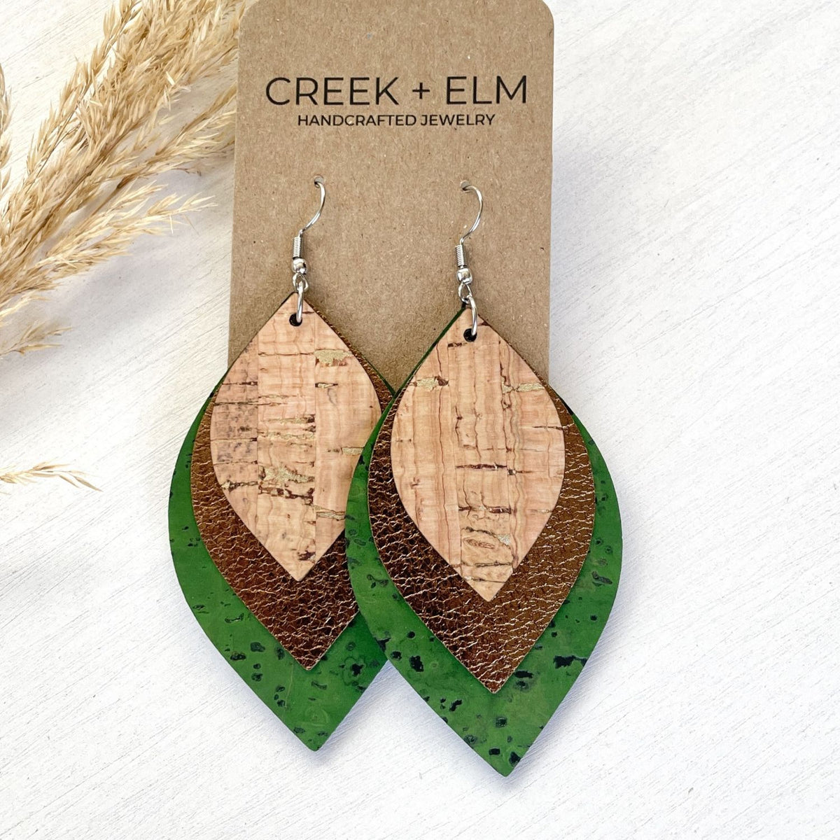 A pair of handmade dangle earrings displayed on a brown kraft card labeled 'CREEK + ELM HANDCRAFTED JEWELRY.' The earrings have silver fishhook wires and layered teardrop-shaped pieces: a light natural wood center, a medium brown textured middle layer, and a dark green speckled outer layer.