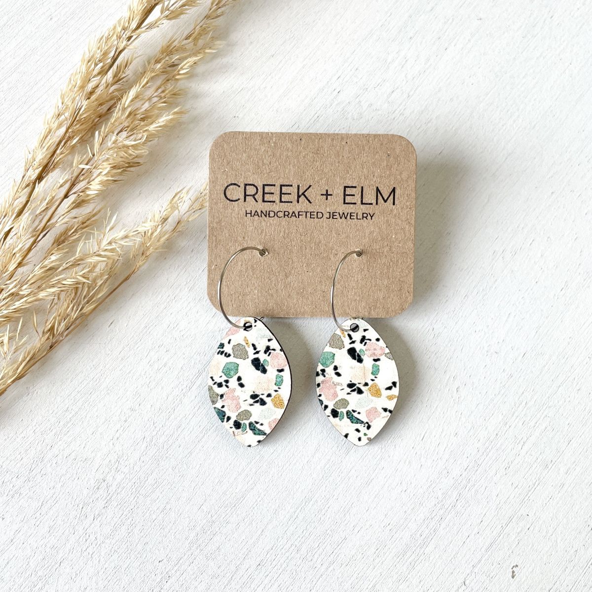 Teardrop-shaped dangling earrings with a terrazzo pattern on a kraft display card, labeled Creek + Elm Handcrafted Jewelry, styled with dried grass on a white textured background.
