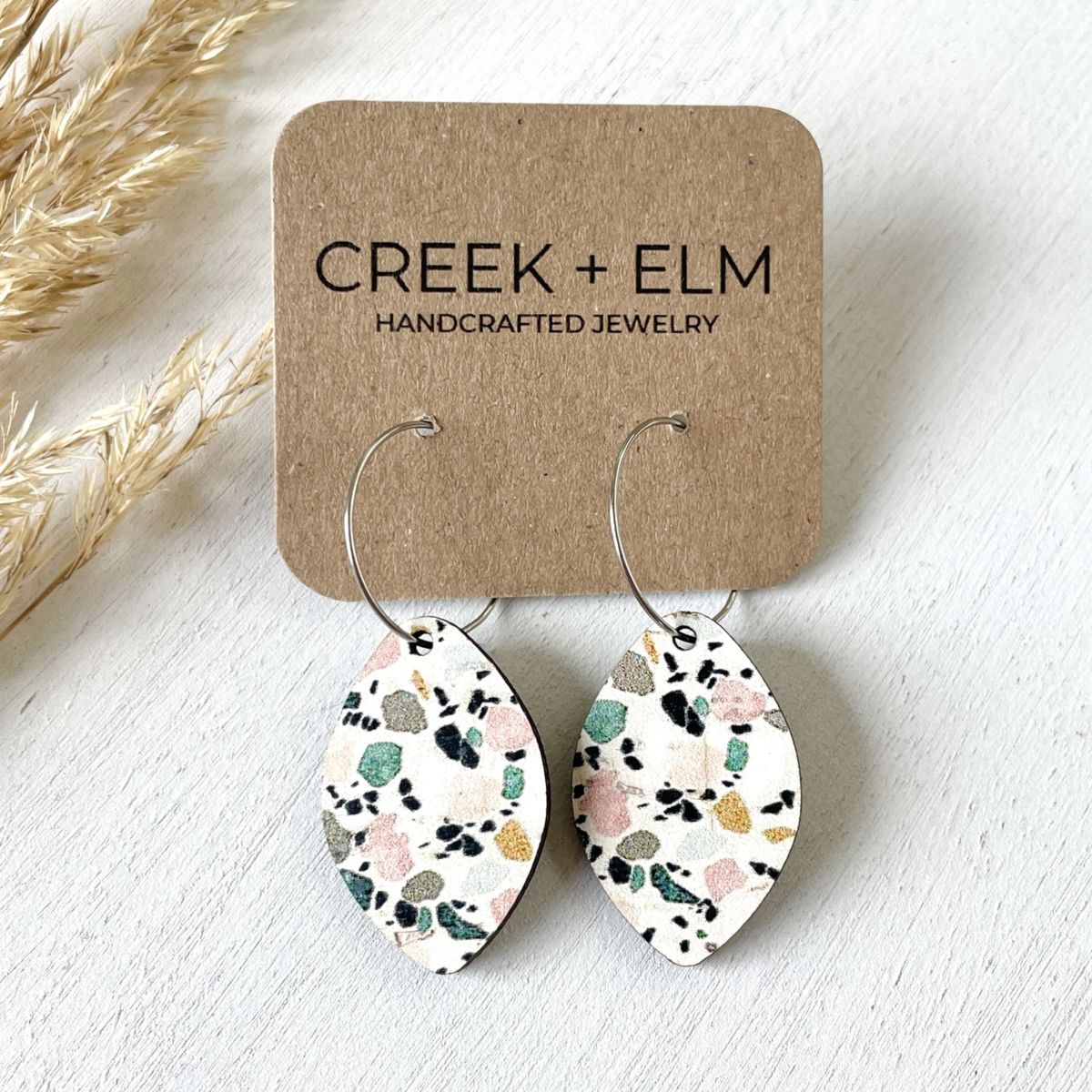 Teardrop-shaped dangling earrings with a terrazzo pattern on a white backing card from Creek + Elm Handcrafted Jewelry, displayed on a light textured surface with dried grass accents.