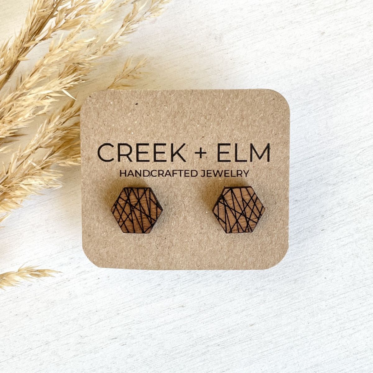 Two hexagonal wooden stud earrings with a laser-cut geometric pattern, mounted on a brown kraft paper display card that reads 'CREEK + ELM HANDCRAFTED JEWELRY'. The card is styled on a white wooden surface next to dried grass.