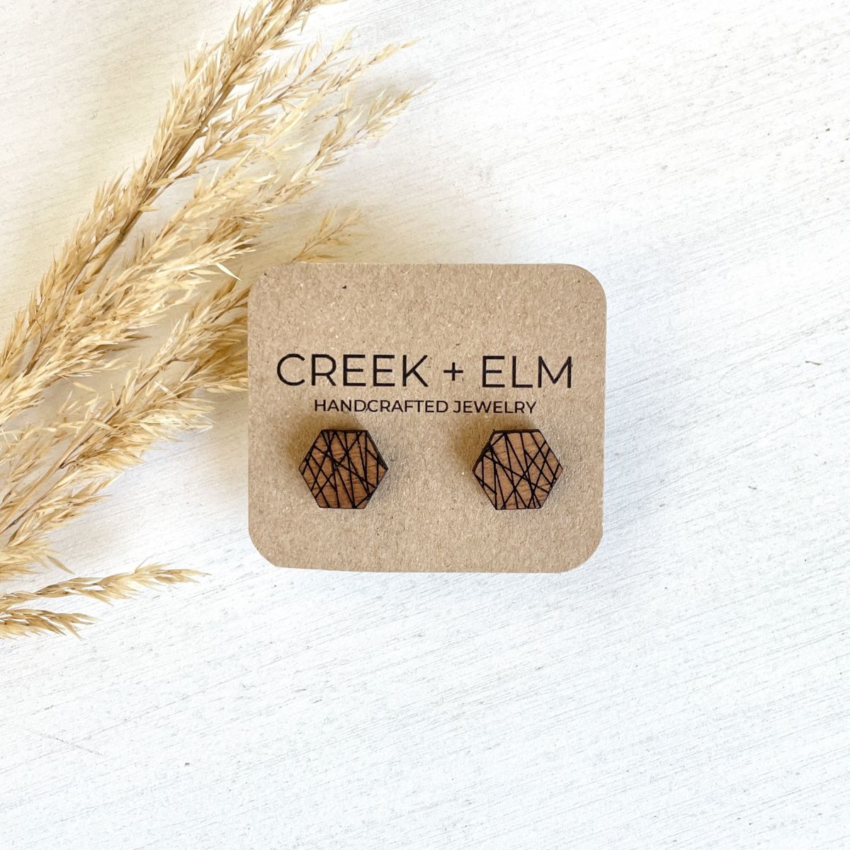 Hexagonal wooden stud earrings with a black laser-engraved geometric pattern, displayed on a brown kraft card labeled 'CREEK + ELM HANDCRAFTED JEWELRY' on a white wooden surface with dried grass.