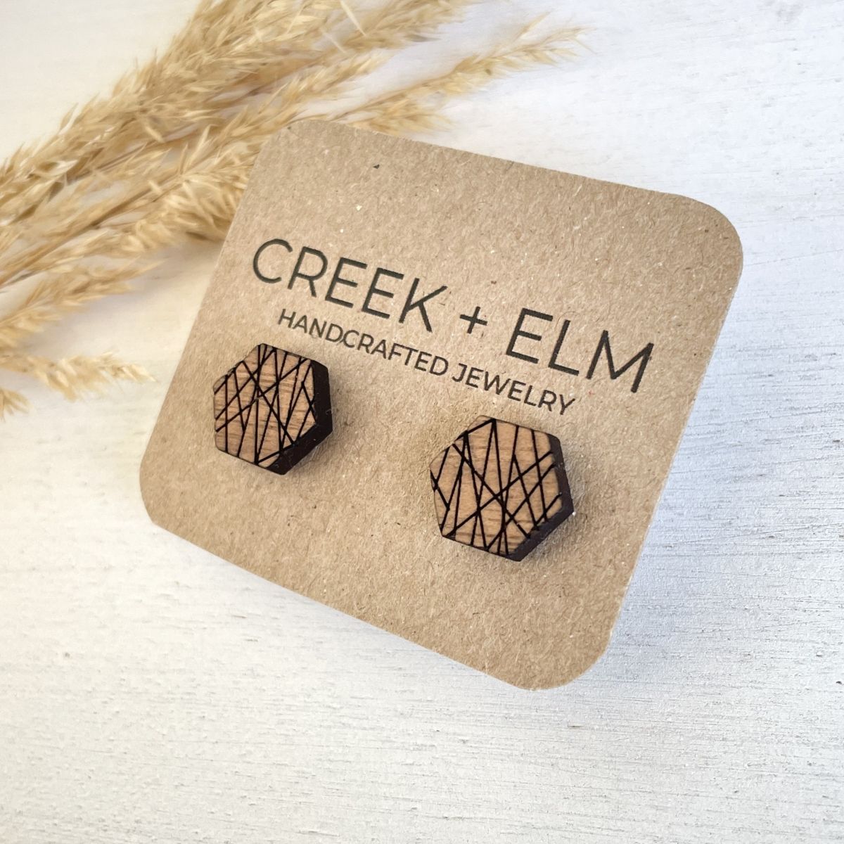 Hexagonal wooden stud earrings with a geometric line pattern, displayed on a brown kraft card labeled 'CREEK + ELM HANDCRAFTED JEWELRY' with dried grass in the background.