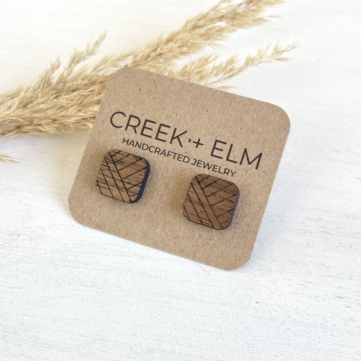 A pair of wooden stud earrings with a geometric basketweave pattern, mounted on a brown kraft display card that reads 'CREEK + ELM HANDCRAFTED JEWELRY'. The card is styled on a white textured surface with dried grass in the background.
