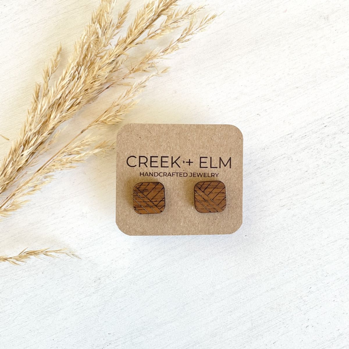 A pair of square wooden stud earrings with a geometric pattern, mounted on a brown kraft display card. The card is placed on a white textured surface next to dried pampas grass.