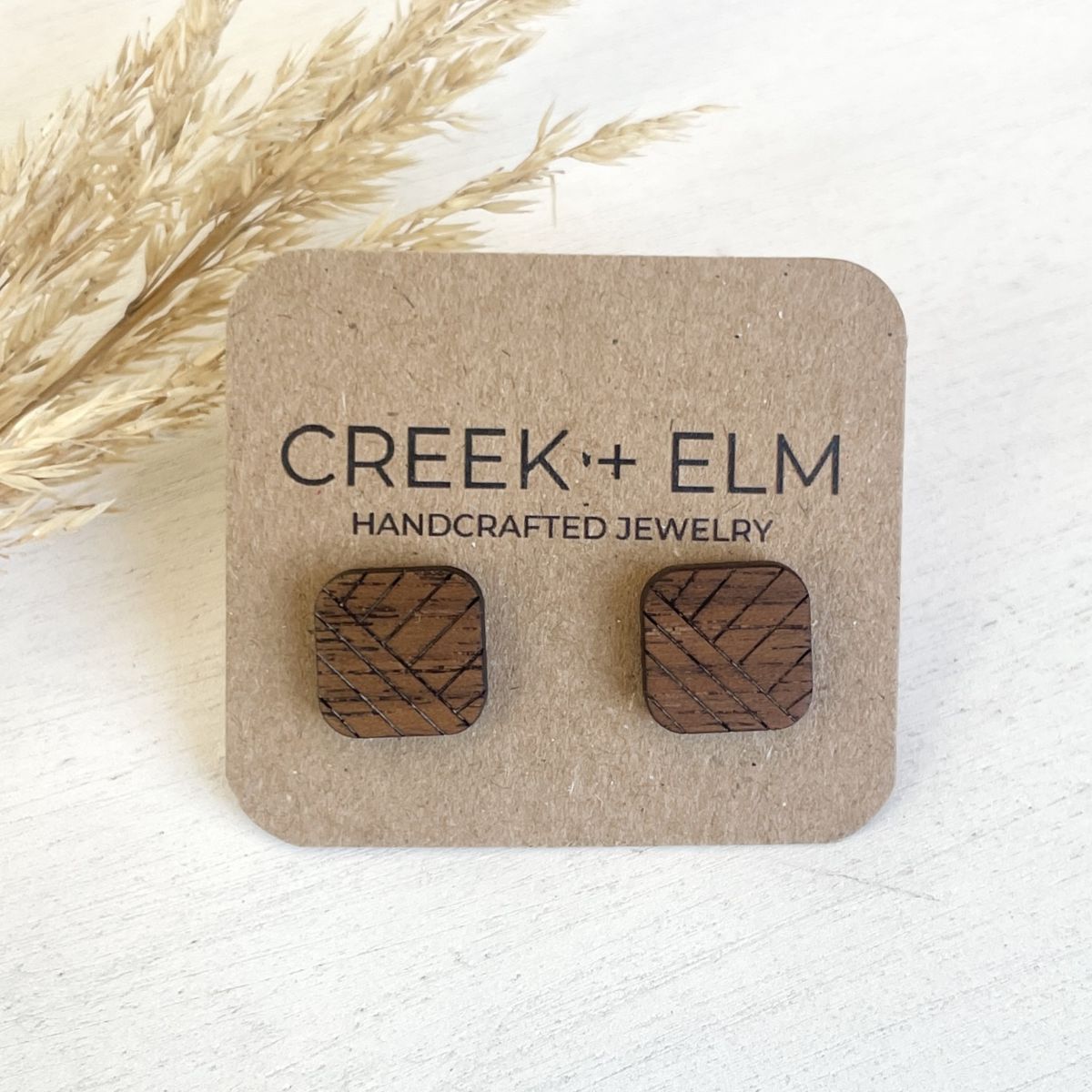 A pair of square wooden stud earrings with a geometric line pattern, mounted on a brown kraft display card that reads 'CREEK + ELM HANDCRAFTED JEWELRY'. The card is styled with dried pampas grass on a light, textured white surface.