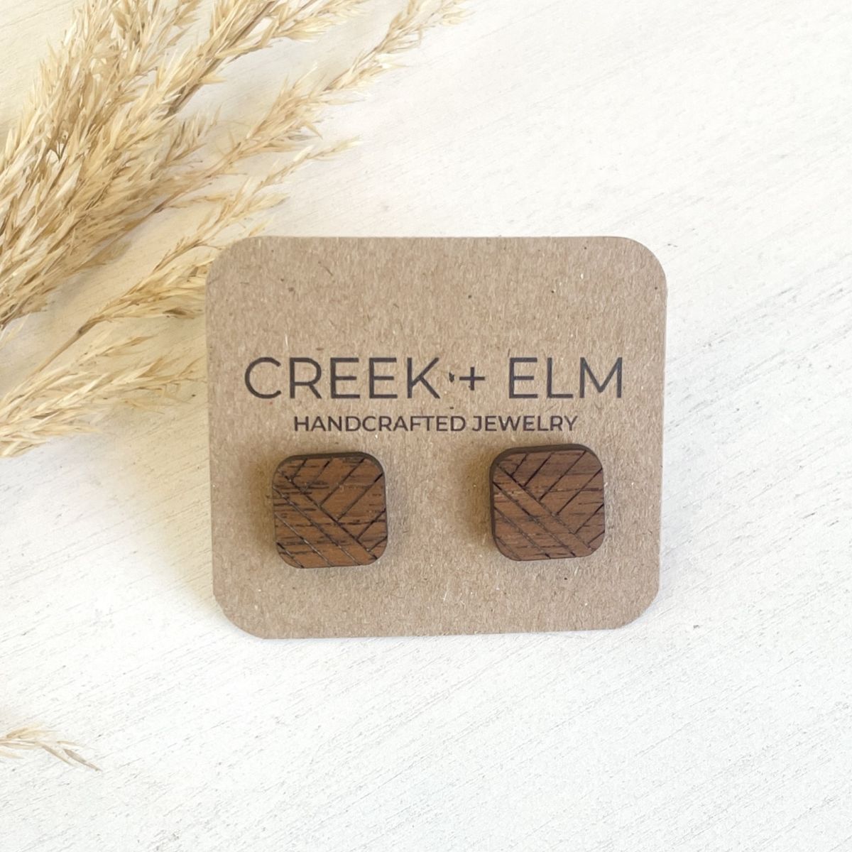 A pair of square wooden stud earrings with a carved geometric pattern, mounted on a brown kraft display card printed with the brand name 'CREEK + ELM'. The card is styled on a white textured surface with dried pampas grass in the upper left corner.